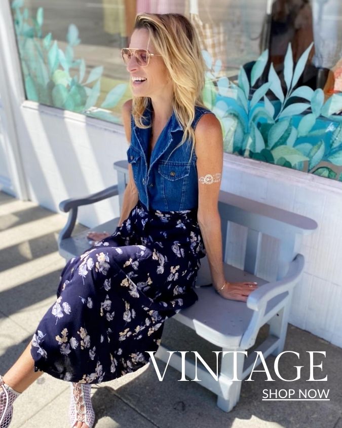 Woman sitting on small bench in front of store wearing denim vest and floral maxi skirt 