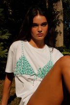 Woman wearing white tee with green bikini top print sitting outdoors in sunlight surrounded by plants