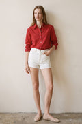 Woman wearing red button-up shirt with rolled sleeves and white high-waisted shorts standing against beige wall