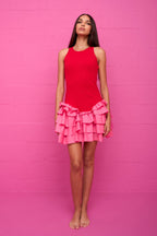 Woman wearing a sleeveless red mini dress with layered pink ruffles standing against a pink wall