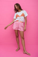Woman wearing a white tee with a textured orange bikini-style top overlay and pink checkered shorts against pink wall