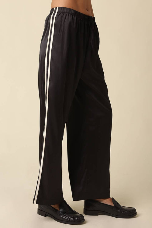 Women's black satin track pants with white side stripes and an elastic waistband styled with black loafers