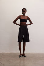 Woman wearing black Bermuda shorts and a strapless black top standing against a plain wall in women's clothing style