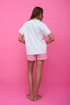 Woman wearing a loose white t-shirt and pink gingham shorts standing barefoot against a bright pink wall