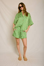 Woman wearing green fish print shorts and matching loose shirt with sunglasses and tan sandals in a neutral studio setting