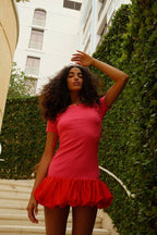 Woman wearing a fitted pink mini dress with a voluminous red bubble hem standing by a green ivy wall outdoors