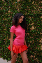 Woman wearing a fitted pink mini dress with a voluminous red bubble hem standing against a leafy wall background