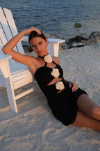 Woman in a black mini dress with cream floral details posing on sandy beach beside white chair by the water