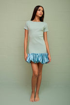 Woman wearing a fitted pastel mini dress with a voluminous satin bubble hem standing against a green wall
