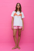 Woman wearing white tee with orange bikini top over it and pink gingham shorts standing against pink wall