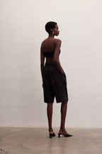 Woman modeling black Bermuda shorts with hands in pockets, wearing black tube top and heels against plain wall