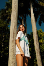 Woman wearing a white T-shirt with a green bikini top design standing among tall palm trees in bright sunlight
