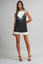 Woman wearing sleeveless mini dress with contrast lace overlay and round neckline, styled with black heels against gray backdrop.