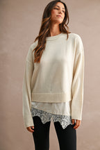 Woman wearing a cream knit sweater with a satin layered hem and floral lace trim over black pants