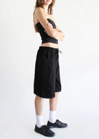 Woman wearing loose black denim jorts with elastic waistband paired with black top and sneakers in casual pose