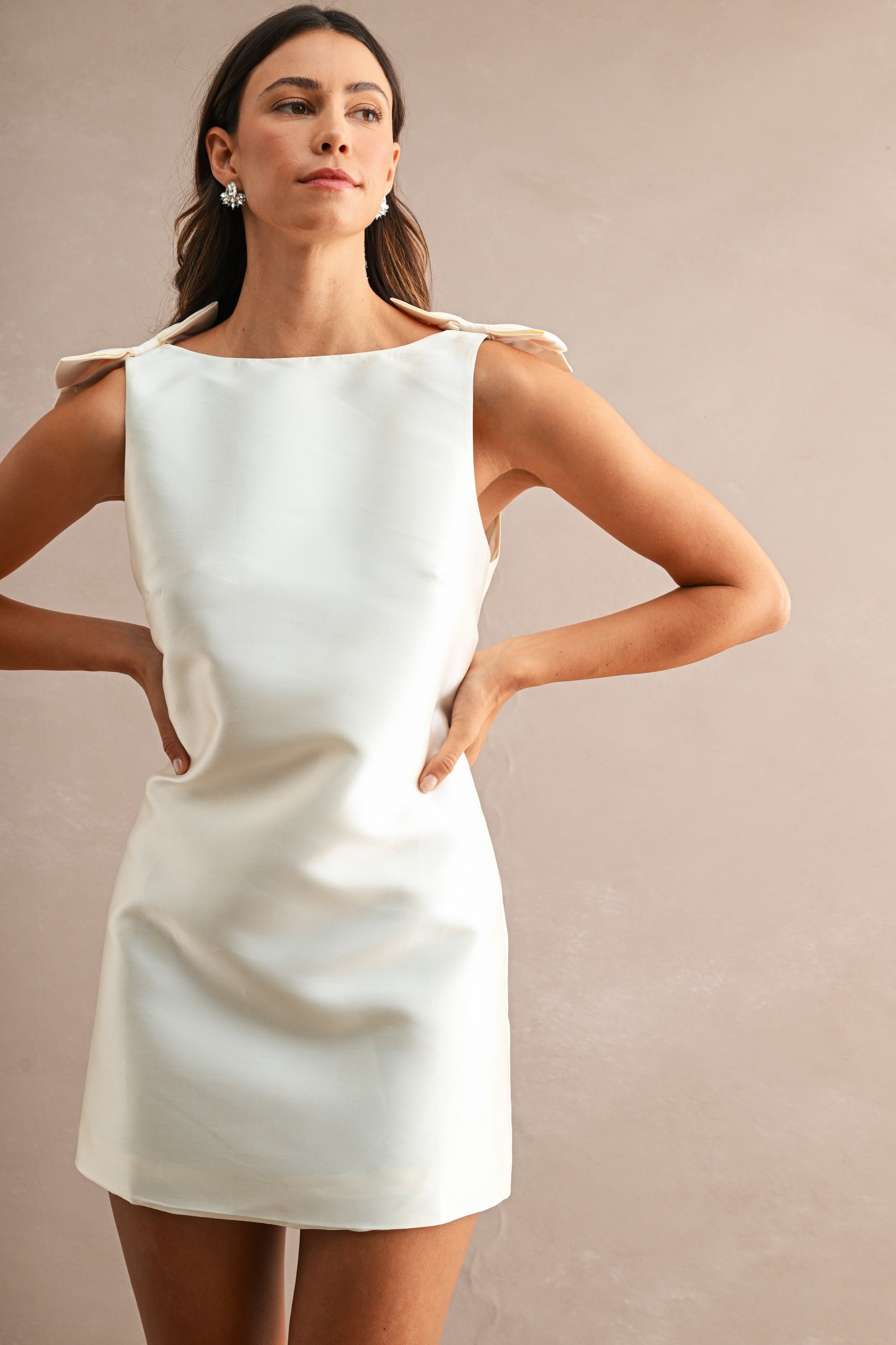 Woman wearing sleeveless white mini dress with bow details on shoulders, posing against beige background.