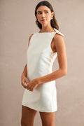 Woman wearing sleeveless white mini dress with boat neckline and bow details on shoulders, stylish women's clothing.