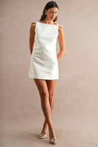 Woman wearing a sleeveless white mini dress with bow details at shoulders and a straight silhouette.