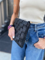 Woman holding black woven vegan leather clutch with detachable strap, wearing white top and blue jeans