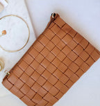 Woven vegan leather clutch in tan with detachable strap, styled on white fabric near gold jewelry and marble tray.