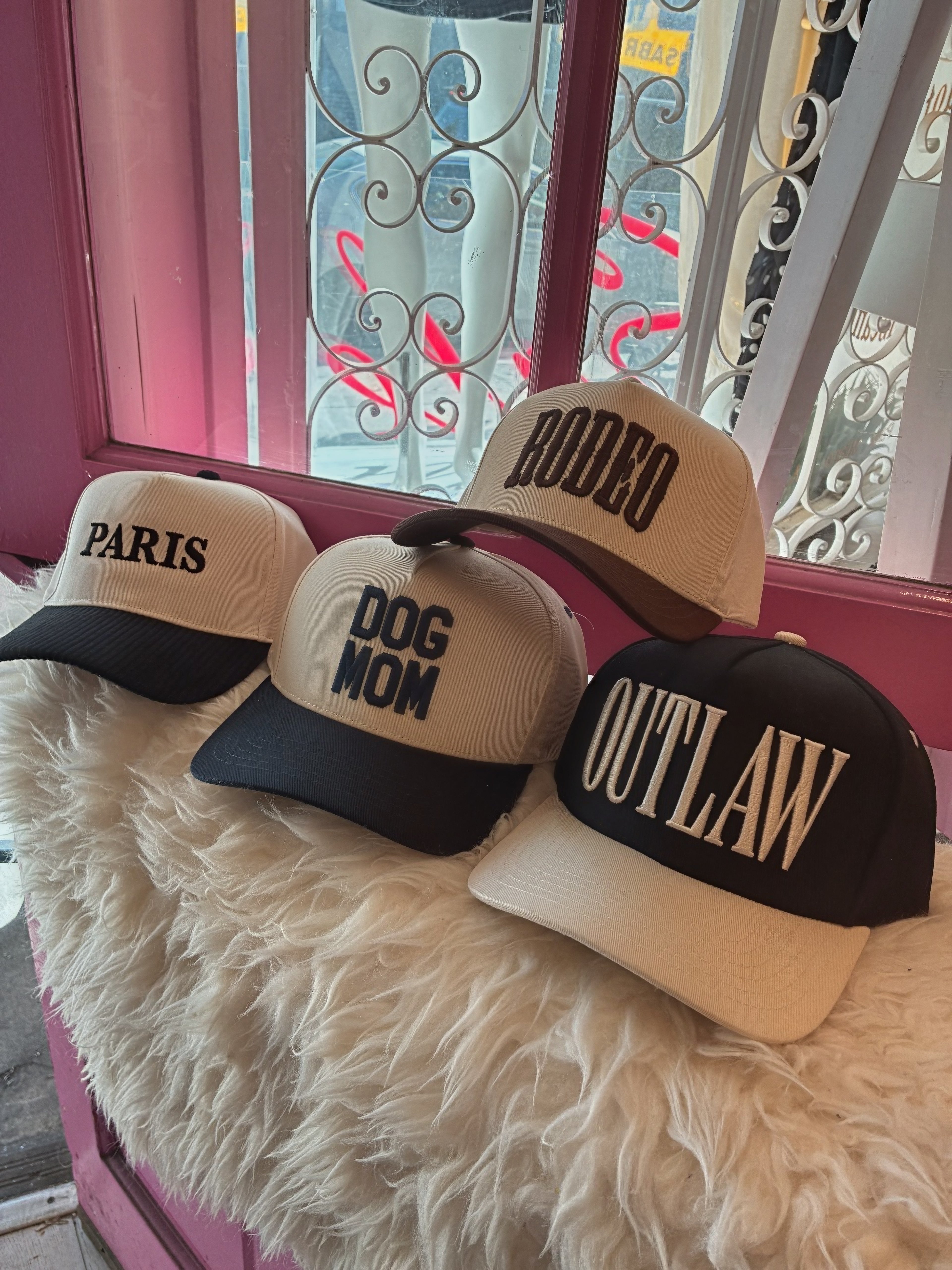 Four two tone baseball hats with bold text designs displayed on a fluffy surface by a window in a fashion store.