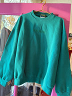 Green vintage-style sweatshirt with embroidered text hanging on gold hanger in women’s clothing store.