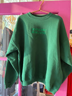 Green women's sweatshirt with "HOPELESS ROMANTIC" embroidered text hanging on a pink door background