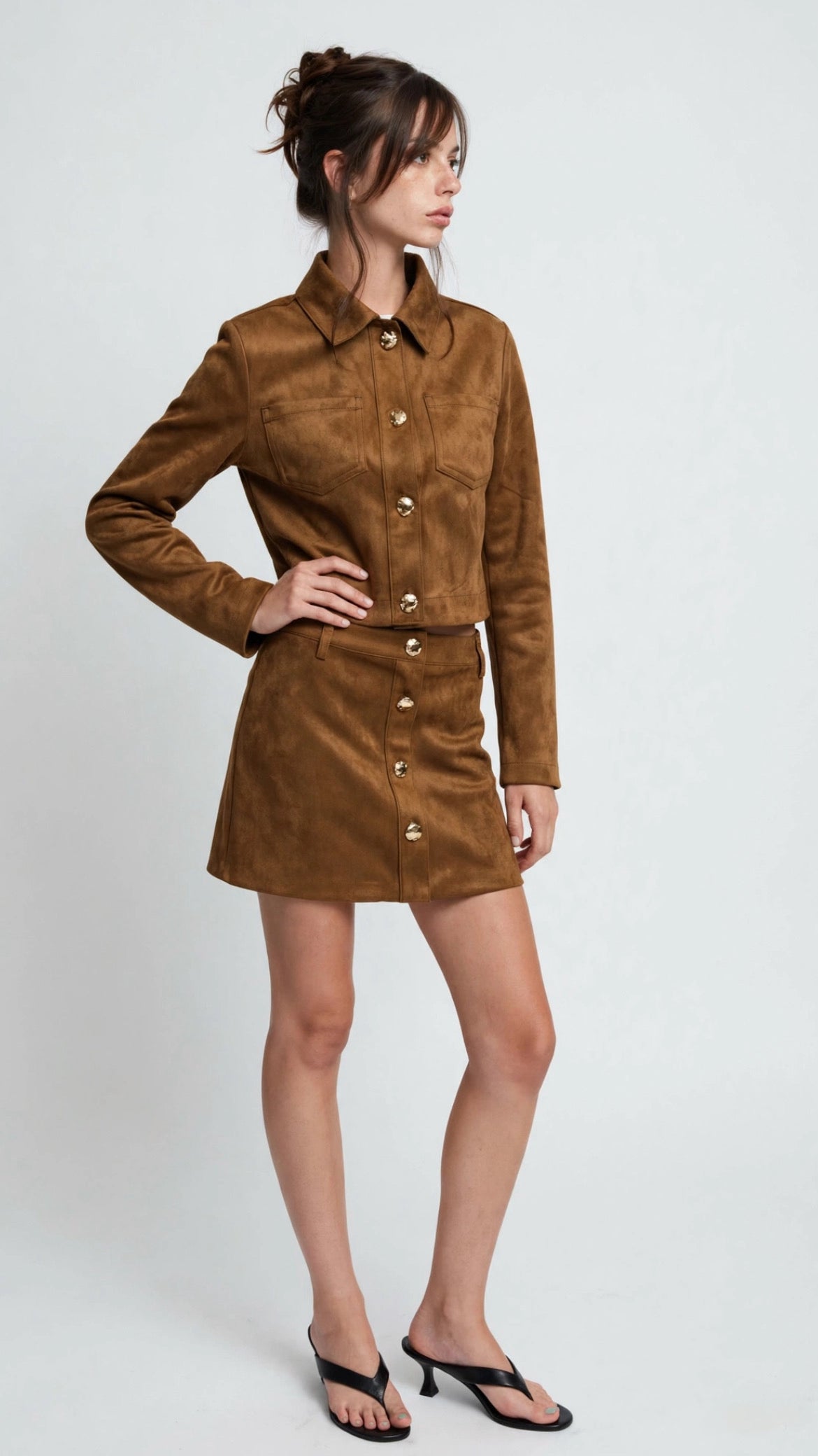 A woman modeling a brown suede long-sleeve shirt dress with a button-down front, collar, and matching belt, paired with black flat sandals, against a light gray studio background.