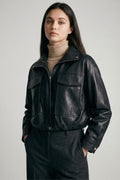 Woman wearing a black cropped vegan leather jacket over a beige turtleneck and dark trousers, hands in pockets