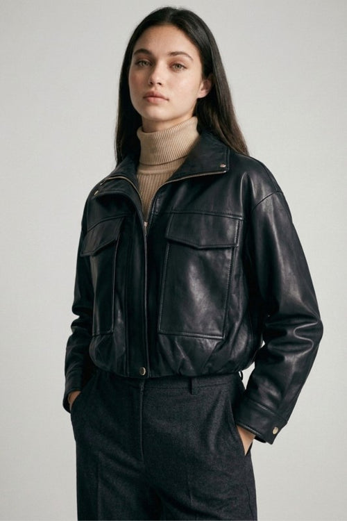 Woman wearing a black cropped vegan leather jacket over a beige turtleneck and dark trousers, hands in pockets