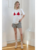 Woman wearing white tee with red bikini graphic and striped shorts in a bright room with wooden floor and columns