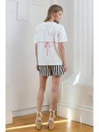Woman wearing white short-sleeve tee with red back tie detail and black-and-white striped shorts in bright room