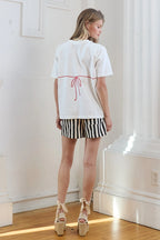 Woman wearing white short-sleeve tee with red bikini-inspired print on back, paired with black and white striped shorts
