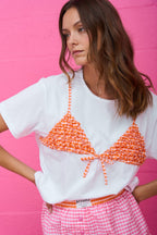 Woman wearing white tee with orange gingham bikini style detail and pink gingham skirt against pink wall