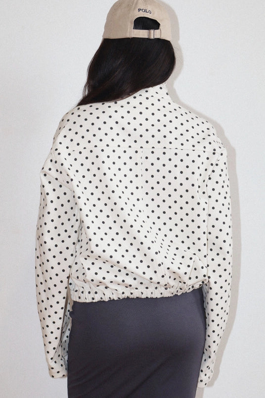 Woman wearing a Polkadots Funnel Jacket with black polka dots on white fabric, paired with a beige cap, viewed from the back.