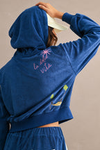 Woman wearing a blue Ciao Terry Hoodie with a hood up, featuring pink "la dolce vita" embroidery and a small palm tree and hammock design on the back.
