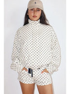 Woman wearing a white Polkadots Funnel Jacket with black polka dots, featuring a high funnel neck and long sleeves, paired with matching shorts and a beige cap.