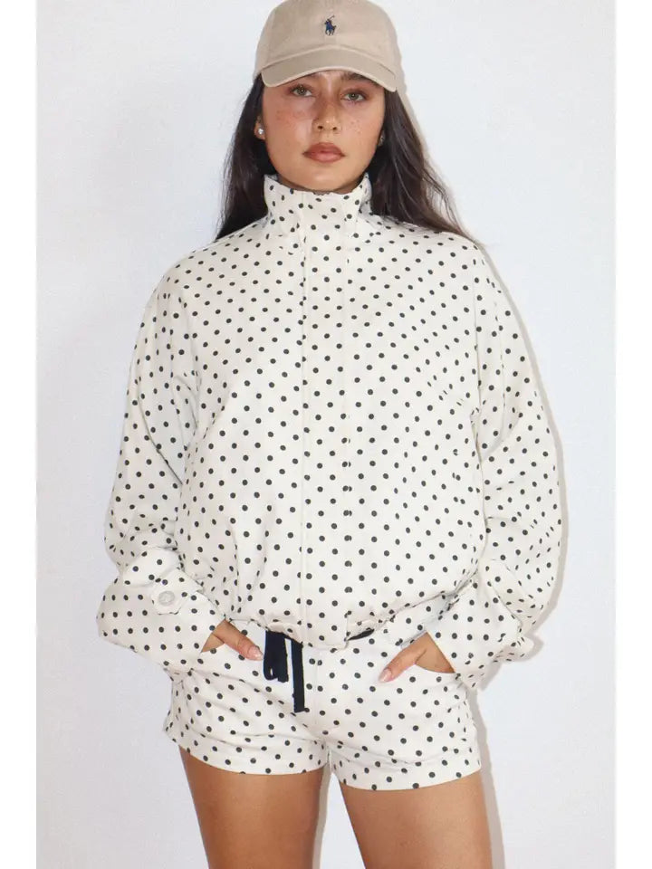 Woman wearing a white Polkadots Funnel Jacket with black polka dots, featuring a high funnel neck and long sleeves, paired with matching shorts and a beige cap.