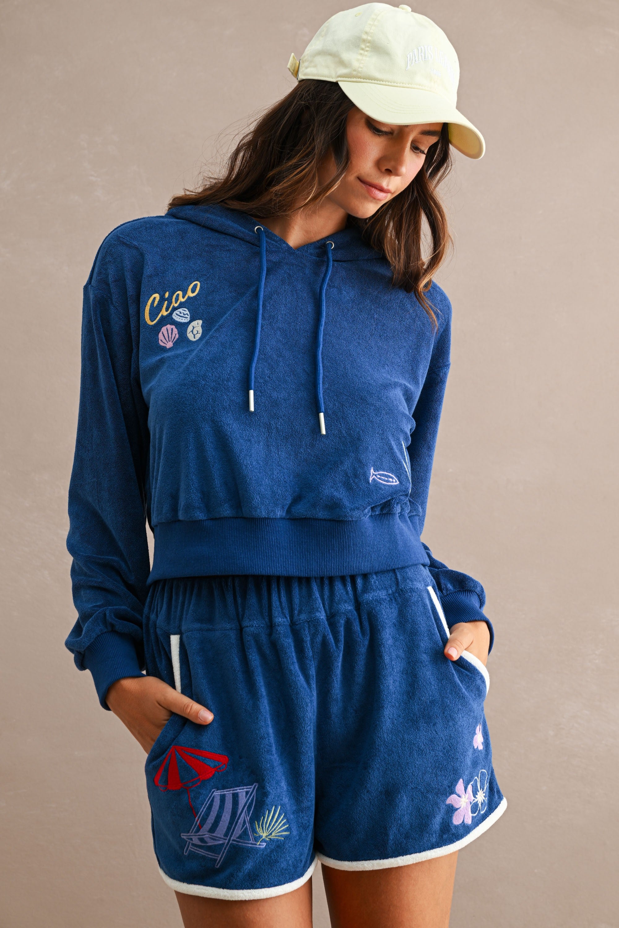 Woman wearing a navy blue Ciao Terry Hoodie with hood and drawstrings, featuring embroidered seashells and fish, paired with matching shorts and a light yellow cap.