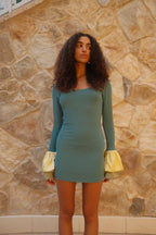 Woman wearing a fitted mini dress with long sleeves and satin ruffle cuffs standing against a stone wall background