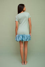 Woman wearing a fitted light blue mini dress with a voluminous satin bubble hem, standing barefoot against a pale wall