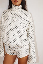 Woman wearing a white Polkadots Funnel Jacket with black polka dots, featuring a high funnel neck and long sleeves, paired with matching shorts.