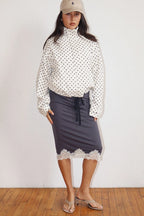 Woman wearing a white Polkadots Funnel Jacket with black polka dots, paired with a dark skirt featuring lace trim at the hem and beige heeled sandals.