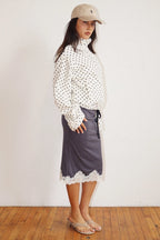 Woman wearing a white polka dot zip-up jacket with a high neck paired with a dark skirt and beige cap and sandals.