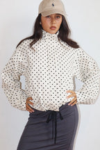 Woman wearing a Polkadots Funnel Jacket with a high collar and black polka dots on a white background, paired with a beige cap and dark skirt, hands on hips.