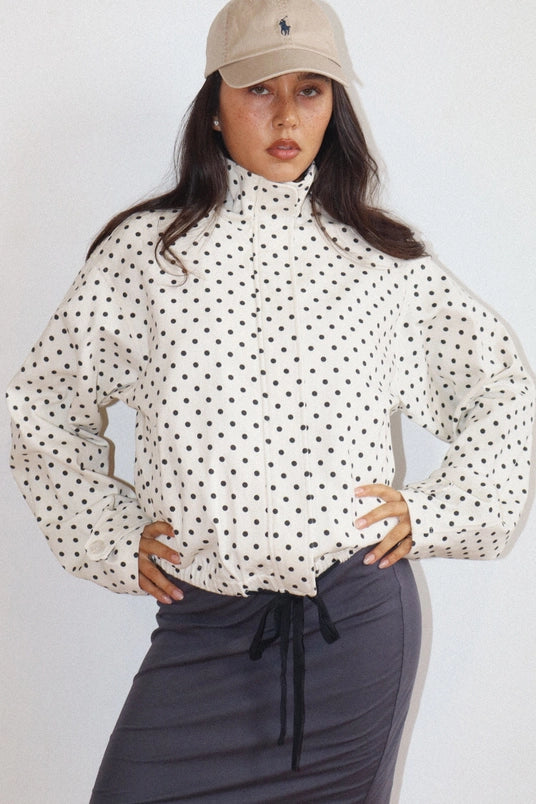 Woman wearing a Polkadots Funnel Jacket with a high collar and black polka dots on a white background, paired with a beige cap and dark skirt, hands on hips.