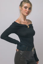 Woman wearing black long-sleeve top with folded neckline and gold buckle, paired with dark pants and belt