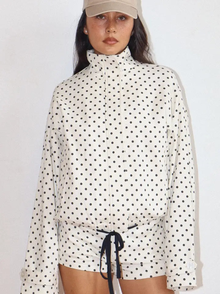 Woman wearing a white Polkadots Funnel Jacket with black polka dots, a high funnel neck, long sleeves, and a black drawstring at the waist, paired with matching shorts.