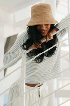 Woman wearing a striped long sleeve top and a woven bucket hat leaning on a white railing indoors