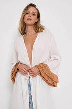 Woman wearing a white kimono with raffia fringed sleeves tied at the waist, styled with jeans.